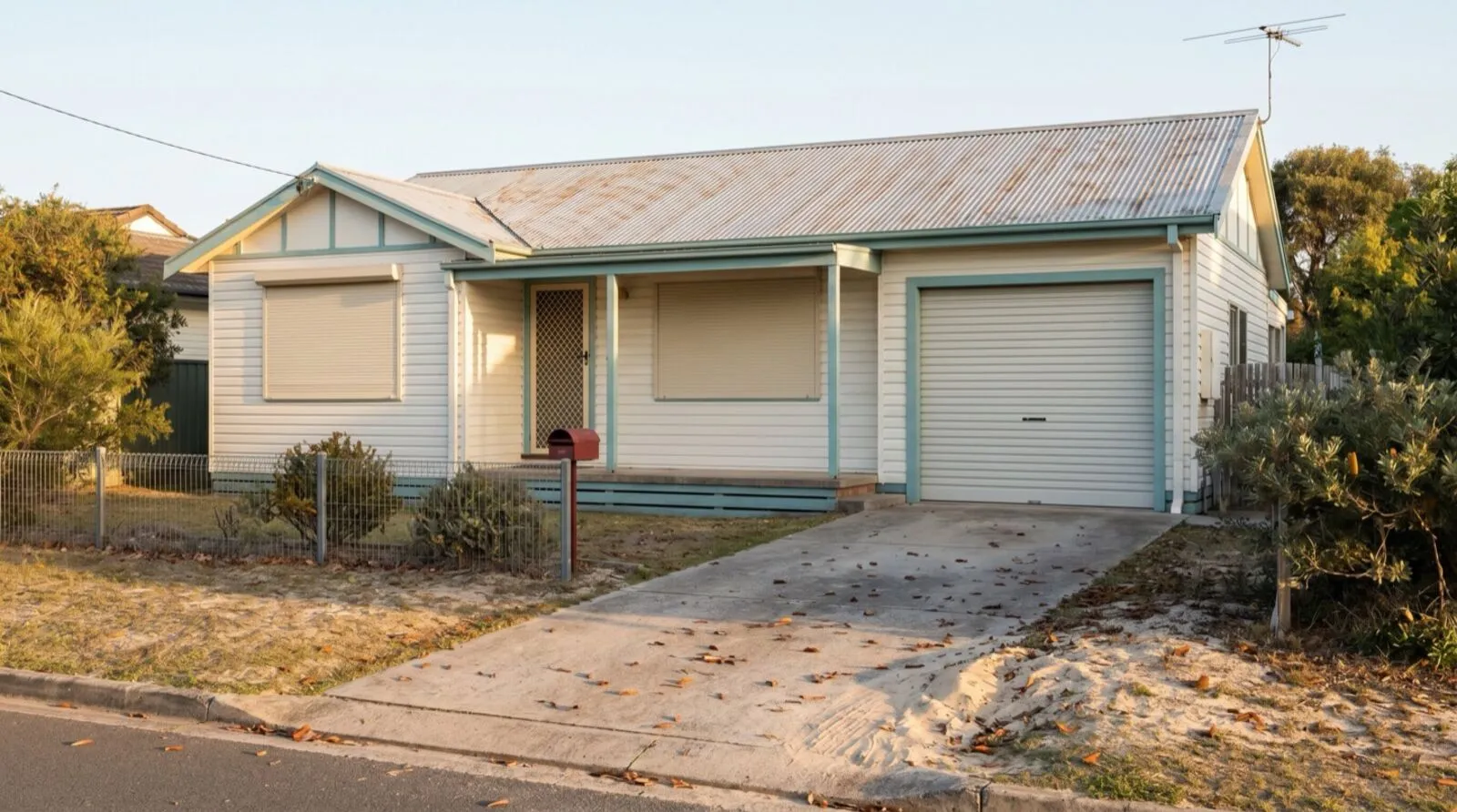 A quiet weatherboard holiday house with a closed roller door and leaves drifted across the empty driveway