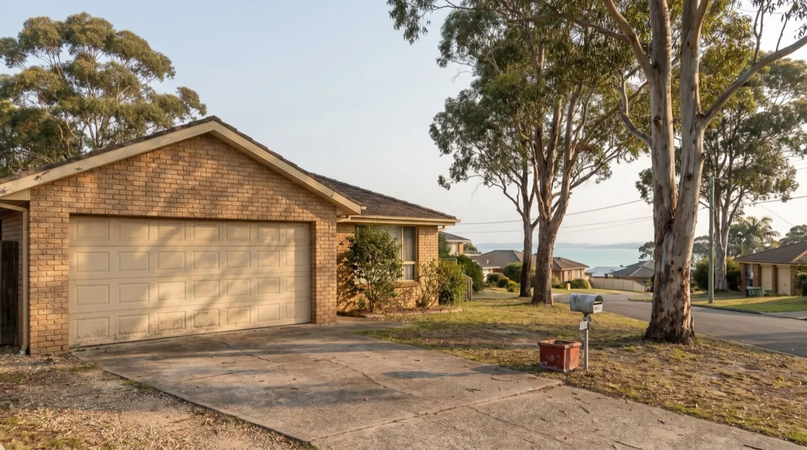 An established brick home in Salamander Bay with a wide sectional garage door, gum trees and a glimpse of the bay